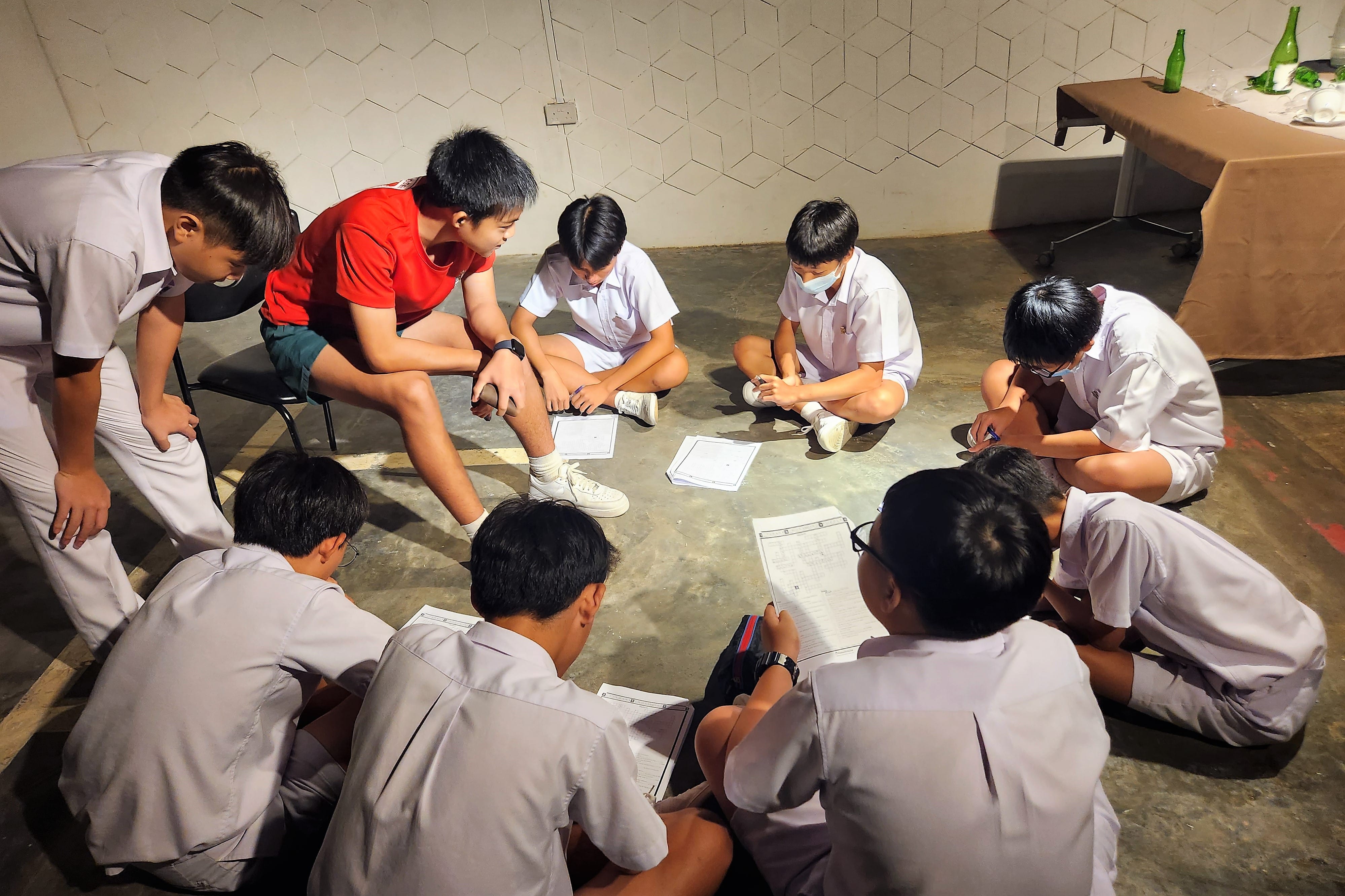A group of students sits in a circle on the floor doing a worksheet activity, with bottles on a table nearby.